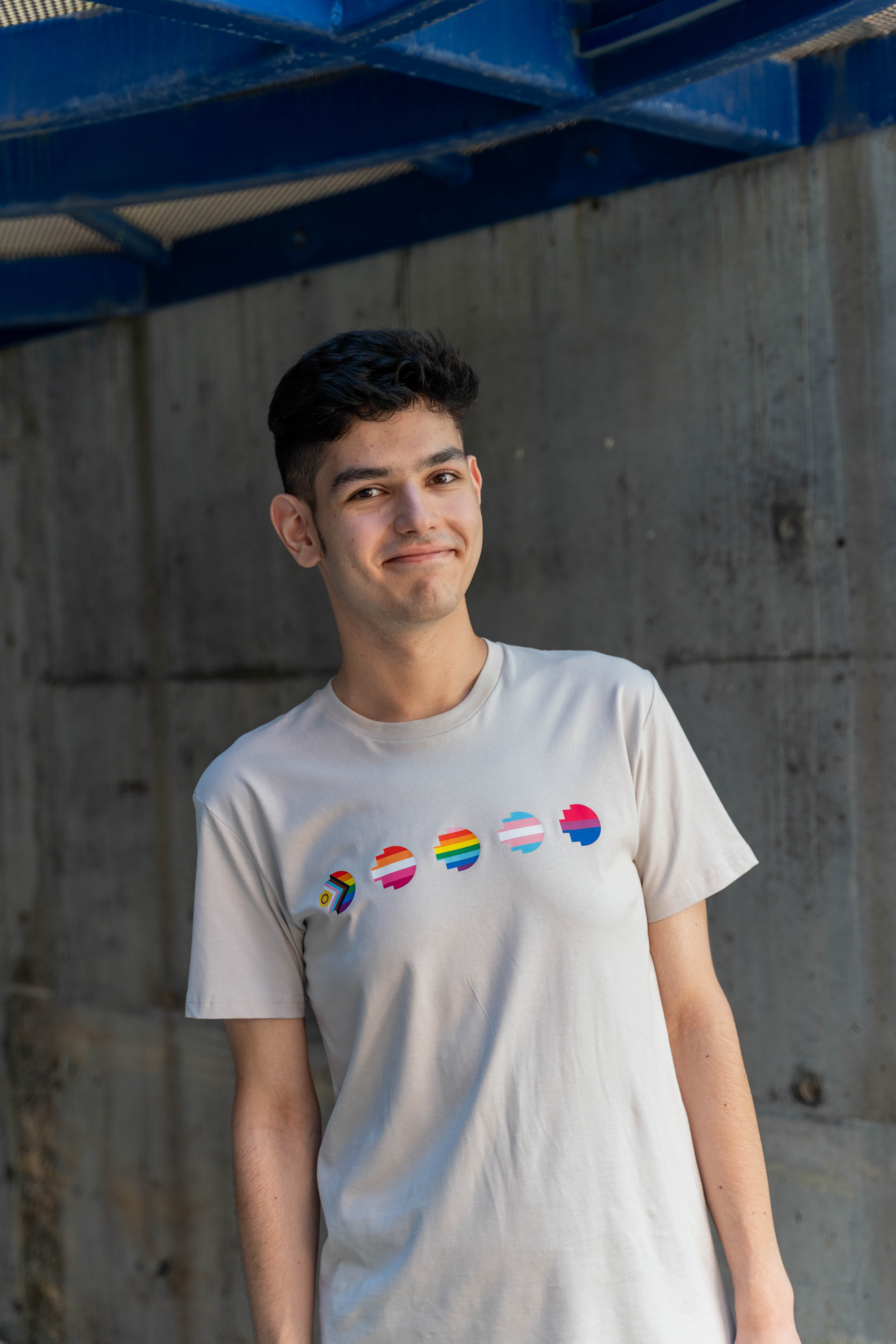RMIT Store Pride Unity Tee in Bone, modelled by a person standing in a casual pose against a neutral background. The soft bone-coloured cotton t-shirt prominently displays five vibrant pride flags across the chest, symbolising inclusivity and unity. The design includes bold, colourful stripes representing different LGBTIQA+ identities. The tee has a relaxed fit, with a round neckline and short sleeves, offering a comfortable, modern style.